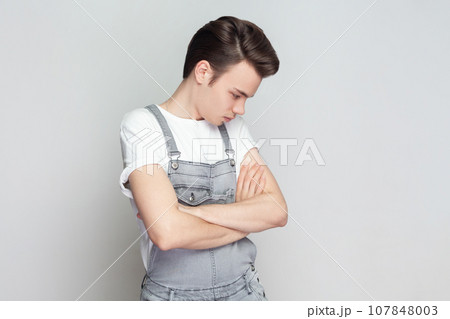 Portrait of sad unhappy young brunette man standing with folded hands, frowns face upset with bad news he received, wearing denim overalls. Indoor studio shot isolated on gray background. 107848003