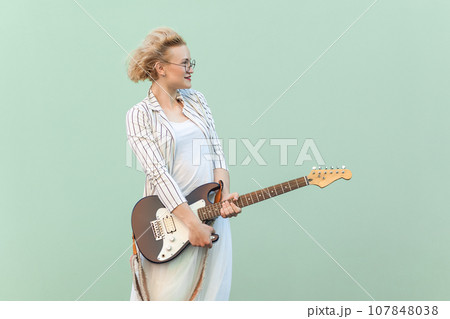 Portrait of beautiful positive blonde woman in white shirt, skirt, and striped blouse with eyeglasses, holding guitar and looking away. Indoor studio shot isolated on light green background. Portrait of beautiful positive blonde woman in white shirt, skirt, and striped blouse with eyeglasses, holding guitar and looking away. Indoor studio shot isolated on light green background. 107848038