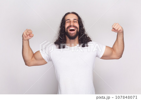 Portrait of happy cheerful strong bodybuilder man in white t-shirt with long wavy hair and beard, standing showing his biceps and triceps. Indoor studio shot isolated on gray background. Portrait of happy cheerful strong bodybuilder man in white t-shirt with long wavy hair and beard, standing showing his biceps and triceps. Indoor studio shot isolated on gray background. 107848071
