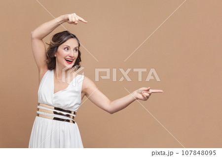 Portrait of amazed excited happy middle aged woman with wavy hair pointing aside at copy space for promotional text, wearing white dress. Indoor studio shot isolated on light brown background. Portrait of amazed excited happy middle aged woman with wavy hair pointing aside at copy space for promotional text, wearing white dress. Indoor studio shot isolated on light brown background. 107848095