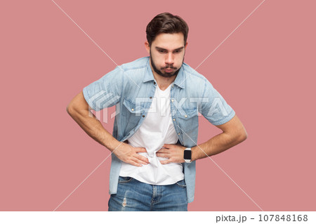 Portrait of bearded man in casual style shirt standing feels stomachache, suffers from indigestion and abdomainal cramps after eating spoiled product. Indoor studio shot isolated on pink background. 107848168