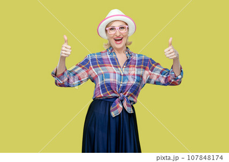 Portrait of senior woman makes thumbs up sign, demonstrates support and respect to someone, smiles pleasantly at camera, achieves desirable goal. Indoor studio shot isolated on yellow background. 107848174