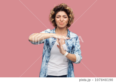 Portrait of sad depressed woman with curly hairstyle wearing blue shirt standing showing time out gesture, looking at camera, expressing sadness. Indoor studio shot isolated on pink background. Portrait of sad depressed woman with curly hairstyle wearing blue shirt standing showing time out gesture, looking at camera, expressing sadness. Indoor studio shot isolated on pink background. 107848190