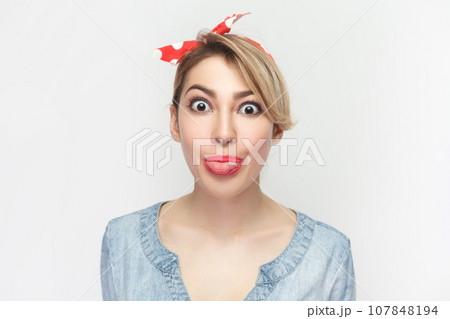 Portrait of funny crazy foolish blonde woman wearing blue denim shirt and red headband standing sticking tongue out, looking at camera. Indoor studio shot isolated on gray background. Portrait of funny crazy foolish blonde woman wearing blue denim shirt and red headband standing sticking tongue out, looking at camera. Indoor studio shot isolated on gray background. 107848194
