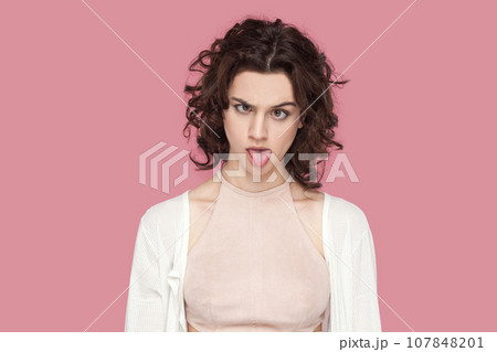 Portrait of crazy fool woman with curly hair wearing casual style outfit having foolish facial expression, grimacing, showing bad manners. Indoor studio shot isolated on pink background. Portrait of crazy fool woman with curly hair wearing casual style outfit having foolish facial expression, grimacing, showing bad manners. Indoor studio shot isolated on pink background. 107848201