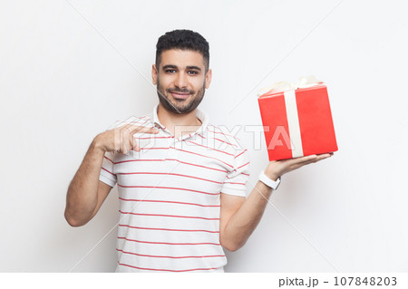 Portrait of smiling positive optimistic bearded man wearing striped t-shirt standing holding red wrapped present box, pointing at gift. Indoor studio shot isolated on gray background. Portrait of smiling positive optimistic bearded man wearing striped t-shirt standing holding red wrapped present box, pointing at gift. Indoor studio shot isolated on gray background. 107848203