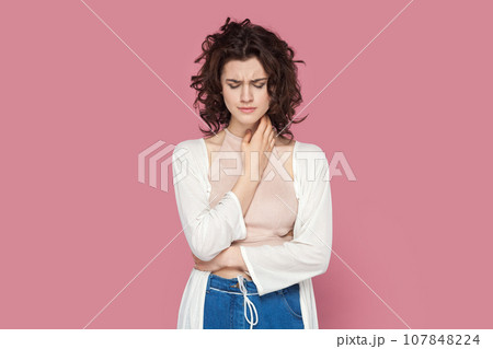 Portrait of sad upset unhappy woman with curly hair wearing casual style outfit standing looking down, crying, hearing bad news. Indoor studio shot isolated on pink background. 107848224