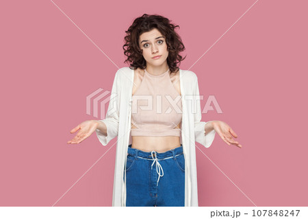 Portrait of puzzled uncertain woman with curly hairstyle wearing casual style outfit spreads hands aside, doesn't know answer. Indoor studio shot isolated on pink background. Portrait of puzzled uncertain woman with curly hairstyle wearing casual style outfit spreads hands aside, doesn't know answer. Indoor studio shot isolated on pink background. 107848247