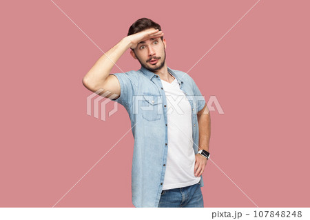 Bearded man in blue casual style shirt standing keeps palm near forehead, looks with concentrated expression into distance, sees something aside. Indoor studio shot isolated on pink background. Bearded man in blue casual style shirt standing keeps palm near forehead, looks with concentrated expression into distance, sees something aside. Indoor studio shot isolated on pink background. 107848248