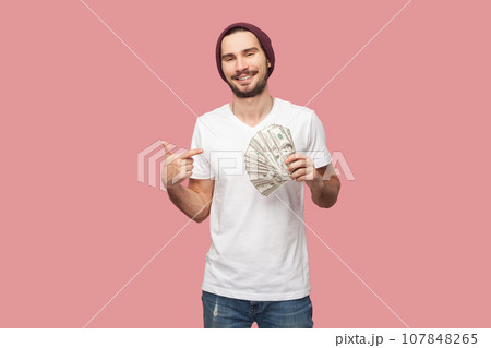 Portrait of satisfied delighted bearded man in white T-shirt and beany hat standing pointing at dollar banknotes, looking smiling at camera. Indoor studio shot isolated on pink background. 107848265