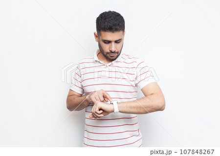 Portrait of concentrated attractive handsome bearded man wearing striped t-shirt standing using his smart watch, uses application. Indoor studio shot isolated on gray background. 107848267