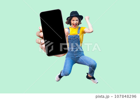 Portrait of happy surprised woman jumping, celebrating her victory, showing big mobile phone with black blank screen, copy space for promotion. Indoor studio shot isolated on light green background. Portrait of happy surprised woman jumping, celebrating her victory, showing big mobile phone with black blank screen, copy space for promotion. Indoor studio shot isolated on light green background. 107848296