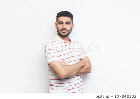 Portrait of delighted positive confident attractive bearded man wearing striped t-shirt standing with crossed hands, looking at camera. Indoor studio shot isolated on gray background. 107848302