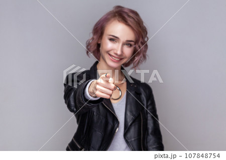 Portrait of smiling satisfied woman with short hairstyle standing pointing fore finger to camera, choosing and needs you, wearing black leather jacket. Indoor studio shot isolated on grey background. 107848754