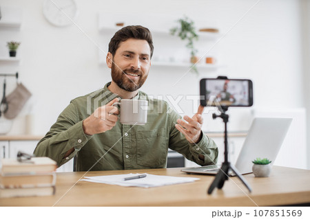 Brunette man recording vlog and holding cup in hand indoor Brunette man recording vlog and holding cup in hand indoor 107851569