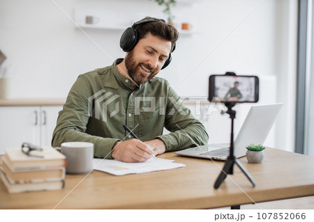 Positive vlogger smiling at mobile camera and making notes Positive vlogger smiling at mobile camera and making notes 107852066