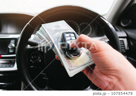 Driver hand holding a car key or keyless remote and usd dollar banknotes in a car Driver hand holding a car key or keyless remote and usd dollar banknotes in a car 107854479