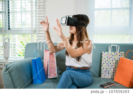 Happy young woman customer wearing 3d VR glasses virtual reality headset with shopping bags around she touching and pointing on air to online shopping in living room at home, digital cyberspace Happy young woman customer wearing 3d VR glasses virtual reality headset with shopping bags around she touching and pointing on air to online shopping in living room at home, digital cyberspace 107856265