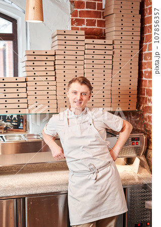 The chef prepares pizza in a neapolitan pizzeria. Cook in a apron in the kitchen with a shovel in his hands. boxes for food delivery on background. The chef prepares pizza in a neapolitan pizzeria. Cook in a apron in the kitchen with a shovel in his hands. boxes for food delivery on background. 107856357