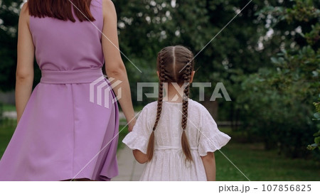 Mother and little daughter walk joining hands along road across city park 107856825
