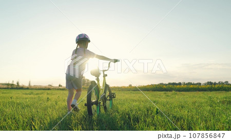 Cute little girl wearing helmet walks pulling bicycle across summer field Cute little girl wearing helmet walks pulling bicycle across summer field 107856847