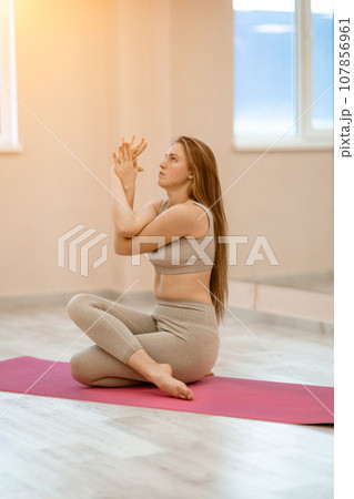 Young woman doing yoga in the gym. A girl with long hair and in a beige tracksuit stands in a cow pose on a pink carpet. A woman performs Gomukhasana. 107856961