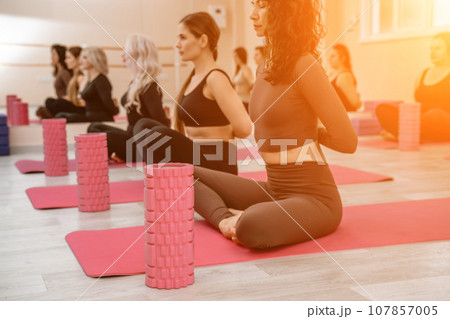 A group of six athletic women doing pilates or yoga on pink mats in front of a window in a beige loft studio interior. Teamwork, good mood and healthy lifestyle concept. A group of six athletic women doing pilates or yoga on pink mats in front of a window in a beige loft studio interior. Teamwork, good mood and healthy lifestyle concept. 107857005
