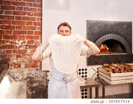 The chef prepare pizza. Raw pizza ready to bake. Cook in a apron in the kitchen Rolls out pizza dough. boxes for food delivery on background. The chef prepare pizza. Raw pizza ready to bake. Cook in a apron in the kitchen Rolls out pizza dough. boxes for food delivery on background. 107857514