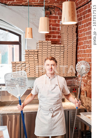 The chef prepares pizza in a neapolitan pizzeria. Cook in a apron in the kitchen with a shovel in his hands. boxes for food delivery on background. 107857515