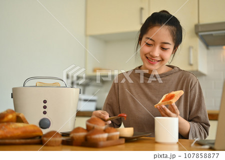 Smiling young woman spreading strawberry jam on slice of bread at kitchen table Smiling young woman spreading strawberry jam on slice of bread at kitchen table 107858877