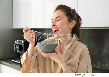 Gorgeous woman starts her day with cereals, eats breakfast and smiles, wears bathrobe, leans on kitchen worktop, starts her morning with quick meal 107860097