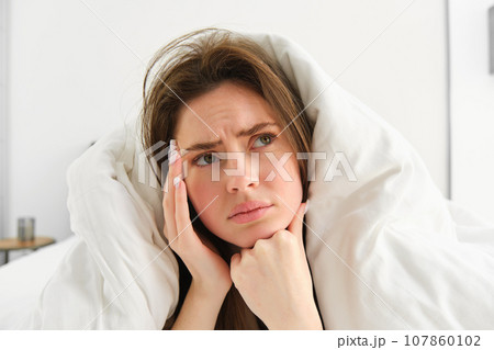 Close up of woman feeling unwell at home, lying in bed under white blankets and frowning, touching head, has headache, migraine, recovering from covid 107860102