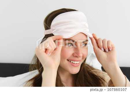 Portrait of smiling young modern woman open her eyes in morning, waking up and taking off sleeping mask, looking outside window, resting in bed, lying in bedroom 107860107