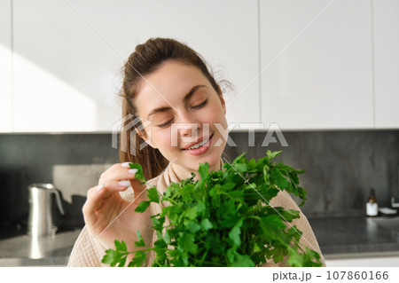 Portrait of beautiful smiling girl with bouquet of parsley, standing in the kitchen and cooking, adding herbs to healthy fresh salad or meal, preparing food 107860166