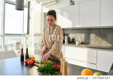 Portrait of woman cooking breakfast, chopping vegetables for salad, using board and knife, standing in the kitchen and wearing bathrobe 107860167