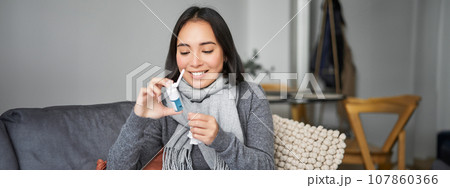 Woman looking at her spray from sore throat, treating her cold, staying at home sick, feeling better after flu, sitting in scarf at home in living room Woman looking at her spray from sore throat, treating her cold, staying at home sick, feeling better after flu, sitting in scarf at home in living room 107860366