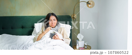 Covid-19, flu and vaccination concept. Korean girl lying in bed with cold, catching influenza, drinking hot tea and using prescribed medication Covid-19, flu and vaccination concept. Korean girl lying in bed with cold, catching influenza, drinking hot tea and using prescribed medication 107860522