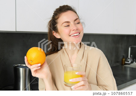 Image of good-looking healthy woman in bathrobe, drinking fresh juice, showing orange fruit, posing in kitchen 107860557