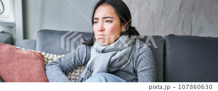 Portrait of sad woman feeling unwell, wearing warm clothes and scarf at her home without heating, staying on sick leave in her house 107860630