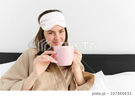 Happy young woman starting her morning with cup of coffee in bed, smiling and enjoying her lazy day, has sleeping mask on forehead Happy young woman starting her morning with cup of coffee in bed, smiling and enjoying her lazy day, has sleeping mask on forehead 107860632