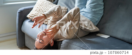 Portrait of beautiful, carefree asian woman lying on sofa, laughing and smiling, listening music in wireless headphones Portrait of beautiful, carefree asian woman lying on sofa, laughing and smiling, listening music in wireless headphones 107860714