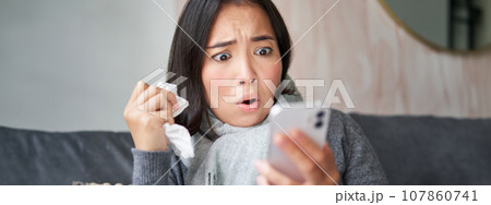 Portrait of ill woman looking at smartphone, looking concerned and shocked at mobile phone, reading bad news while sitting on sick leave at home Portrait of ill woman looking at smartphone, looking concerned and shocked at mobile phone, reading bad news while sitting on sick leave at home 107860741