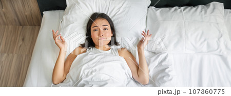 Portrait of asian girl lying in her bed and shrugs, looks puzzled, overthinking before going to sleep, looking complicated at camera 107860775