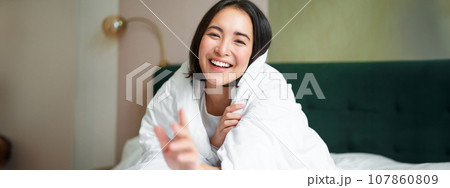 Happy beautiful asian woman covered with blanket, sits on bed in warm duvet, tries to reach camera with a hand, smiling playfully. Hotel bedroom, comfort and apartments concept Happy beautiful asian woman covered with blanket, sits on bed in warm duvet, tries to reach camera with a hand, smiling playfully. Hotel bedroom, comfort and apartments concept 107860809