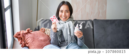 Portrait of happy smiling asian girl showing medication, sore throat spray and drugs from flu or cold, staying at home, wearing warm clothes Portrait of happy smiling asian girl showing medication, sore throat spray and drugs from flu or cold, staying at home, wearing warm clothes 107860815