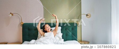 Portrait of smiling happy asian girl, wakes up feeling enthusiastic, stretches her hands up, enjoys good morning in her bedroom Portrait of smiling happy asian girl, wakes up feeling enthusiastic, stretches her hands up, enjoys good morning in her bedroom 107860821