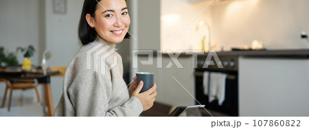 Portrait of smiling asian girl with cup of coffee, drinking hot tea and getting cozy at home. Warming up with beverage 107860822