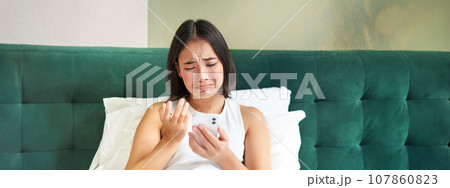 Portrait of sad frowning korean girl, lying in bed and looking at smartphone with upset, disappointed face expression, using mobile phone, complaining Portrait of sad frowning korean girl, lying in bed and looking at smartphone with upset, disappointed face expression, using mobile phone, complaining 107860823