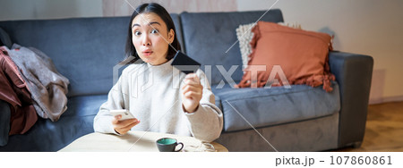 Young woman in living room, showing her credit card and using smartphone to pay, order online or shopping on application Young woman in living room, showing her credit card and using smartphone to pay, order online or shopping on application 107860861