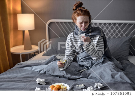 young woman with cold blowing runny nose. female sneezing in tissue in bedroom 107861095